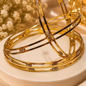 Op de afbeelding: Een set van drie goudkleurige metalen armbanden met een dubbelbandontwerp, verbonden door kleine, heldere, sprankelende steentjes die gelijkmatig rond de omtrek van elke cirkelvormige armband zijn geplaatst voor een glanzend effect.