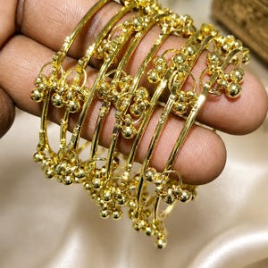 May include: A set of six gold-colored bangles, each adorned with small, spherical embellishments. The bangles are held in a hand, showcasing their intricate design and polished finish. The background is a soft, neutral color.