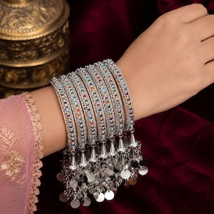 May include: A set of six silver-toned metal bangles featuring a filigree pattern inlaid with small, multicolored circular stones. Each bangle is adorned with a dangling silver-toned charm element consisting of a small cone and flat circular discs.