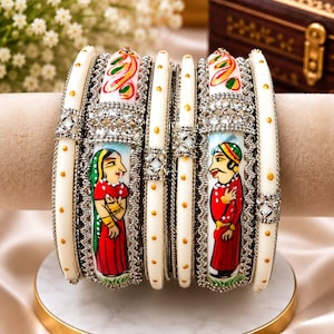 Op de afbeelding: Een set witte armbanden met gouden stippen en zilverkleurige metalen accenten. Twee armbanden tonen handgeschilderde figuren van een persoon in een rood kledingstuk met een groene hoofdbedekking en een persoon in een rode tuniek met een groen-gouden tulband.