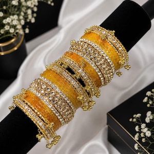 May include: A stack of yellow bangles featuring clear rhinestone embellishments and small gold-toned metal charms. The bracelets are arranged in a cylindrical formation, showcasing a textured surface and a metallic finish on the edges.