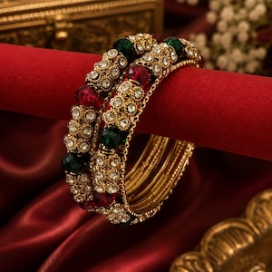 May include: A set of gold-toned bangles featuring intricate metalwork embedded with clear white rhinestones, deep red faceted beads, and dark green faceted beads. The bangles have a textured, ornate surface design and a polished metallic finish.