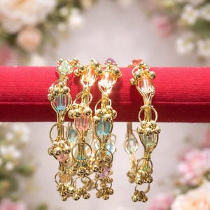 May include: Four gold-toned metal bangles featuring a delicate wire design with small gold spheres and translucent pastel-colored beads in shades of pink, blue, and green. The bracelets are arranged in a row, showcasing a repeating pattern of metallic loops and beads.