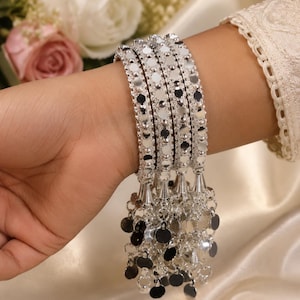 May include: A set of four silver-toned bangles featuring a reflective mirror-like surface and a decorative border. Each bangle is adorned with a dangling cluster of small, circular silver-toned metallic discs that hang from the bottom edge.