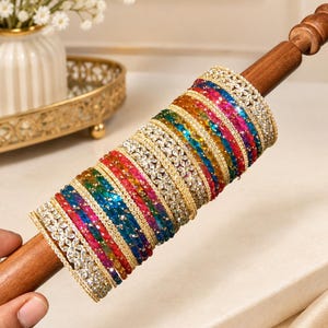 Puede incluir: Un conjunto de brazaletes multicolores dispuestos en una varilla de madera. La colección incluye pulseras en tonos rojo, azul, verde y amarillo, intercaladas con brazaletes dorados con piedras claras y brillantes en un patrón floral.