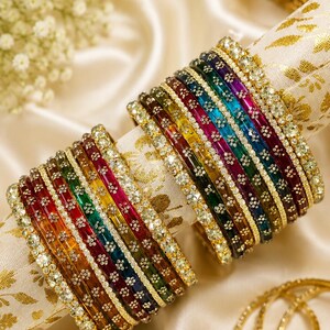 Può includere: Una collezione di braccialetti colorati con un arcobaleno di tonalità, tra cui rosso, arancione, giallo, verde, blu e viola. Ogni braccialetto è adornato con dettagli scintillanti e posto su un tessuto dorato e crema.