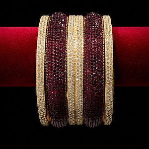 May include: A set of gold-toned bangles with alternating bands of deep red and clear, sparkling stones. The bangles are displayed on a red velvet surface against a black background, showcasing the jewelry's intricate design and color contrast.