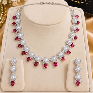 May include: A silver-toned necklace and earring set featuring round white pearls surrounded by clear crystals and accented with oval-shaped red stones. The necklace has a chain with a lobster clasp, and the earrings are drop-style with three pearls.