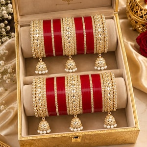 Op de afbeelding: Een set van twee rode en gouden armbanden in een gouden doos. Elke set heeft brede rode banden met gedetailleerd goudfiligraan, heldere stenen en kleine bungelende klokvormige bedels met piepkleine witte kraaltjes aan de onderkant.