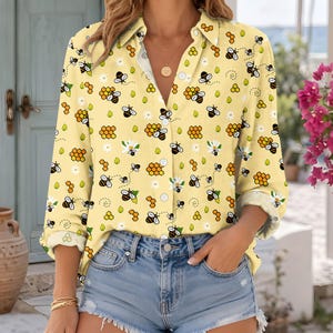 May include: A light yellow long-sleeve button-down shirt featuring an all-over print of small black and yellow bees, orange honeycomb clusters, and white daisy flowers. The shirt has a classic collar, white buttons, and long sleeves.