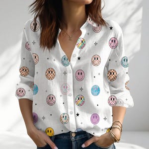 May include: White button-down shirt featuring a repeating pattern of smiley faces in various colours and designs, including checkered and tie-dye patterns. Black star shapes are scattered throughout. The shirt has long sleeves and a collared neckline.