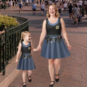 May include: Two sleeveless dresses featuring a police uniform graphic print. The design includes a black bodice with a gold badge emblem, a blue skirt, and a printed utility belt with pouches and a radio. The dresses have a scoop neckline.