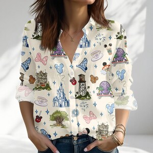 May include: A long-sleeve button-down shirt in an off-white color featuring an all-over print of various theme park icons, including castles, water towers, teacups, monorails, mouse-ear balloons, bows, and star shapes in blue, pink, and brown.