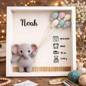Op de afbeelding: Een handgemaakt aandenken voor de kinderkamer met een gehaakte grijze olifant die pastelkleurige ballonnen vasthoudt. De lijst bevat de naam "Noah", geboortedatum, tijd, lengte (48 cm) en gewicht (2.410 g).
