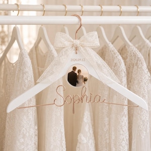 May include: A white wooden clothes hanger with a rose gold metal hook and wire script reading Sophia. A circular photo of a couple is centred on the hanger, topped with a cream lace bow and the printed date 2026.09.12 on the white frame.