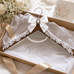 Peut inclure: Un cintre en bois foncé orné d'une bordure en dentelle florale blanche, d'un nœud en satin blanc avec une perle centrale et d'un collier de perles blanches. Le fil métallique forme le prénom Mia, un cœur et la date 12.03.2027.