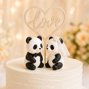 Op de afbeelding: Een taarttopper met twee pandabeeldjes op een witte taart. De ene panda draagt een zwarte vlinderdas, de andere een witte bloem en een doorschijnende sluier. Achter de panda's staat een houten hartvormige uitsparing met het woord love erin.