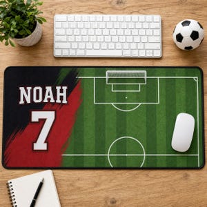 以下が含まれることがあります： 緑色のサッカーフィールドのグラフィック、白いライン、ゴールが描かれた長方形のデスクマット。左側には赤と黒のブラシストロークデザインがあり、白い文字でNOAH、太字のスポーツフォントで数字の7がプリントされています。