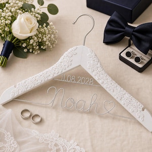 May include: A white wooden clothes hanger decorated with white floral lace trim. The hanger features silver wire bent into the name Noah, a heart shape, and the date 11.08.2028. The hanger includes a silver metal hook at the top center.