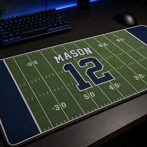May include: A rectangular desk mat featuring a green football field design with white yard lines and numbers. The name MASON is printed in white capital letters above the large dark blue number 12. The mat has dark blue borders on both ends.