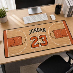 Puede incluir: Una alfombrilla de escritorio rectangular con diseño de cancha de baloncesto, patrón de madera clara, líneas blancas y bordes naranjas. El texto JORDAN está impreso en negro sobre el número 23 grande de color naranja con borde negro.