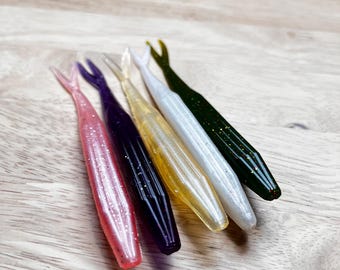 Könnte beinhalten: Fünf Weichplastik-Angelköder in einer Reihe, jeder in einer anderen Farbe. Die Farben sind Rosa, Lila, Gelb, Weiß und Grün. Die Köder liegen auf einer hellbraunen Holzoberfläche.