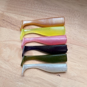 May include: A collection of six soft plastic fishing lures in various colors, including brown, yellow, pink, black, green, and white. Each lure has a paddle tail design and is arranged on a wooden surface.