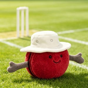May include: A plush, round, red character with a textured surface, two grey arms, and a stitched smiling face. The character wears a white bucket hat with small metal eyelets. The plush toy is positioned on a green surface with white lines.