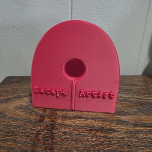 May include: A bright red, 3D-printed object with the words "Escape Artist" embossed on the front. The object has a rounded top with a circular hole in the center and a rectangular base.