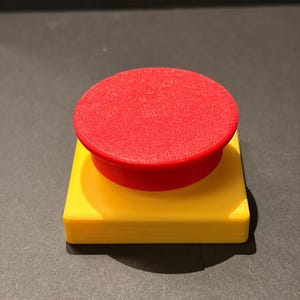 May include: A 3D-printed push button with a bright red circular top and a yellow square base. The button is a solid color and has a smooth texture. The button is likely a component for electronics or a similar device.