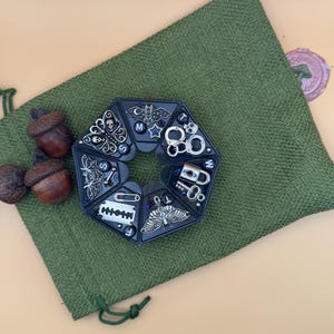 May include: A black hexagonal organizer case containing various silver-toned metal charms, including a skull, butterfly, bat, razor blade, safety pin, moth, and handcuffs. The case is positioned on a textured green fabric pouch with two brown acorns.