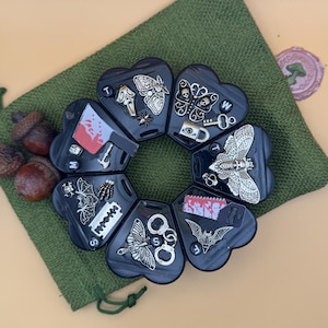 May include: A black heart-shaped weekly pill organizer with seven compartments, each labeled with a day of the week and decorated with silver-toned charms including moths, skulls, a cleaver, a razor blade, handcuffs, and bats.