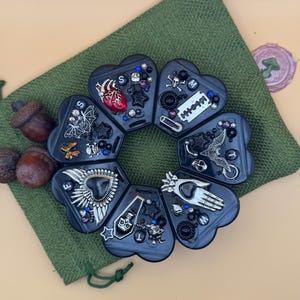 May include: A circular seven-compartment black plastic pill organizer with heart-shaped sections. Each section is decorated with various charms including a red anatomical heart, bats, skulls, a razor blade, a coffin, and a hand, labeled with letters.