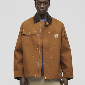 May include: A brown canvas chore coat featuring a dark brown corduroy collar, a front metal zipper closure, two chest pockets with flaps, two lower patch pockets, and a small white Carhartt logo patch on the left chest pocket.