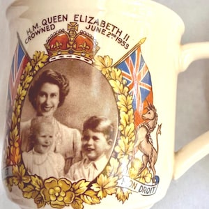 May include: Cream-colored ceramic mug commemorating the coronation of H.M. Queen Elizabeth II on June 2nd, 1953. Features a portrait of the Queen with two children, a crown, flags, and the words "H.M. Queen Elizabeth II Crowned June 2nd 1953".