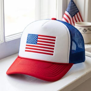 Könnte beinhalten: Eine Trucker-Cap mit weißem Vorderteil und einem aufgestickten amerikanischen Flaggen-Patch. Die Kappe hat einen einfarbig roten Schirm mit einer dünnen roten Kordel an der Basis des Vorderteils. Die Seiten- und Rückenteile bestehen aus blauem Netzstoff.