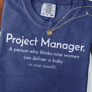 May include: A blue short-sleeved t-shirt with white text that reads: Project Manager. A person who thinks nine women can deliver a baby in one month. The shirt features a Comfort Colors brand label at the inner neckline and a gold-toned necklace.