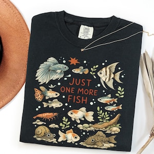 May include: A black crewneck t-shirt featuring a colorful graphic of various fish, a shrimp, and a snail. The text "JUST ONE MORE FISH" is printed in orange in the center. The design includes aquatic plants, bubbles, and a small orange star.
