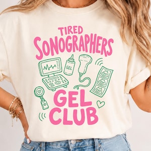 May include: A cream-colored t-shirt featuring the pink text TIRED SONOGRAPHERS GEL CLUB and green line art illustrations of an ultrasound machine, a bottle of gel, a transducer probe, a heart-shaped badge reel, and a strip of three ultrasound images.
