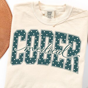 May include: A cream-colored t-shirt featuring the text CODER in large, dark teal block letters with white polka dots. The word medical is written in a cursive, dark teal script across the center. The shirt has a Comfort Colors brand label.