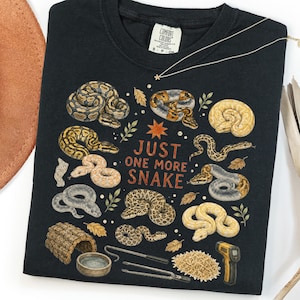 May include: A black crew neck t-shirt featuring a graphic print of various snake species, small leaves, and the text "JUST ONE MORE SNAKE" in orange. The design includes a snake hide, a water bowl, metal tongs, and a digital temperature gun.