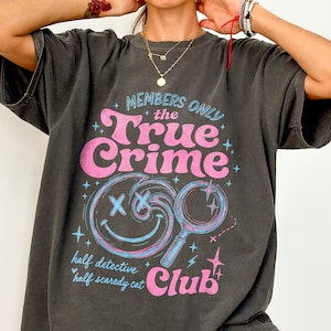 May include: A charcoal gray short-sleeve t-shirt featuring a graphic print with the text "MEMBERS ONLY the True Crime Club" and "half detective half scaredy cat". The design includes a pink smiley face with X eyes and a blue magnifying glass.