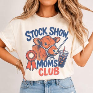 Puede incluir: Una camiseta de color crema con un gráfico de una vaca marrón con cabestro, una escarapela roja y un vaso de plástico con pajita. El texto STOCK SHOW MOMS CLUB aparece en letras azules y rojas en la parte frontal de la prenda.