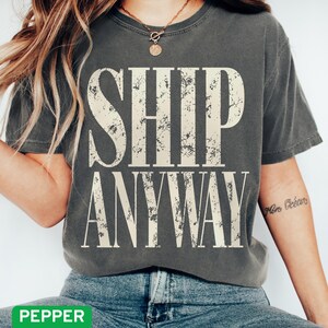 May include: A charcoal gray short-sleeve t-shirt featuring the text SHIP ANYWAY in a large, distressed, off-white serif font centered on the chest. The garment has a crew neckline and a relaxed fit, showcasing a worn-in texture.