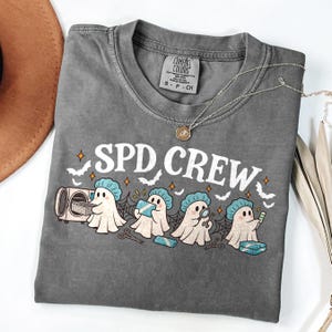 May include: A charcoal gray t-shirt featuring the text SPD CREW in white letters. The graphic shows four ghosts wearing blue surgical caps, interacting with medical instruments, a sterilization unit, and scissors against a spiderweb background.