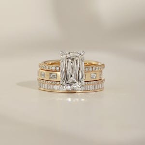 May include: A stack of three gold rings featuring a large rectangular radiant-cut diamond centre stone in a silver-toned setting. The stack includes a thin pavé diamond band, a band with bezel-set baguette diamonds, and a channel-set baguette diamond band.