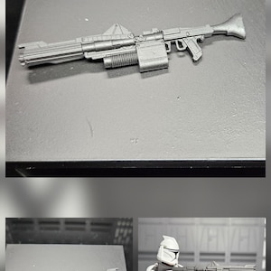 May include: A gray plastic toy blaster rifle with a long barrel, a rectangular magazine, and a flared stock. The weapon features a top-mounted sight and a ribbed cylindrical component beneath the barrel. A white and black armored figure holds the rifle.