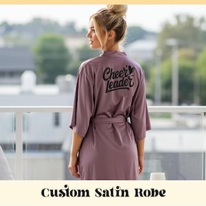 Peut inclure: Une robe en satin mauve avec le texte "Cheer Leader" imprimé dans le dos en écriture noire. La robe a une ceinture assortie et arrive au genou. L'image comprend également le texte "Custom Satin Robe".