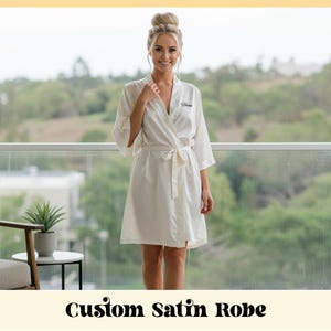 Puede incluir: Una bata de satén color crema con cinturón a juego, usada por una persona en un balcón. La bata tiene un nombre personalizado bordado en la parte delantera. El texto "Custom Satin Robe" se muestra en la parte inferior de la imagen.
