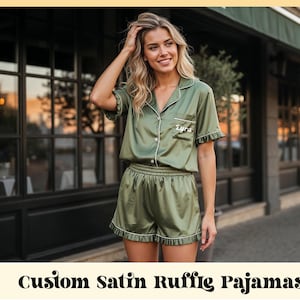 May include: Olive green satin pajama set with white trim. The short-sleeved top has a breast pocket with the name "Lyra" embroidered on it. The shorts have a ruffled hem. The text "Custom Satin Ruffle Pajamas" is at the bottom of the image.